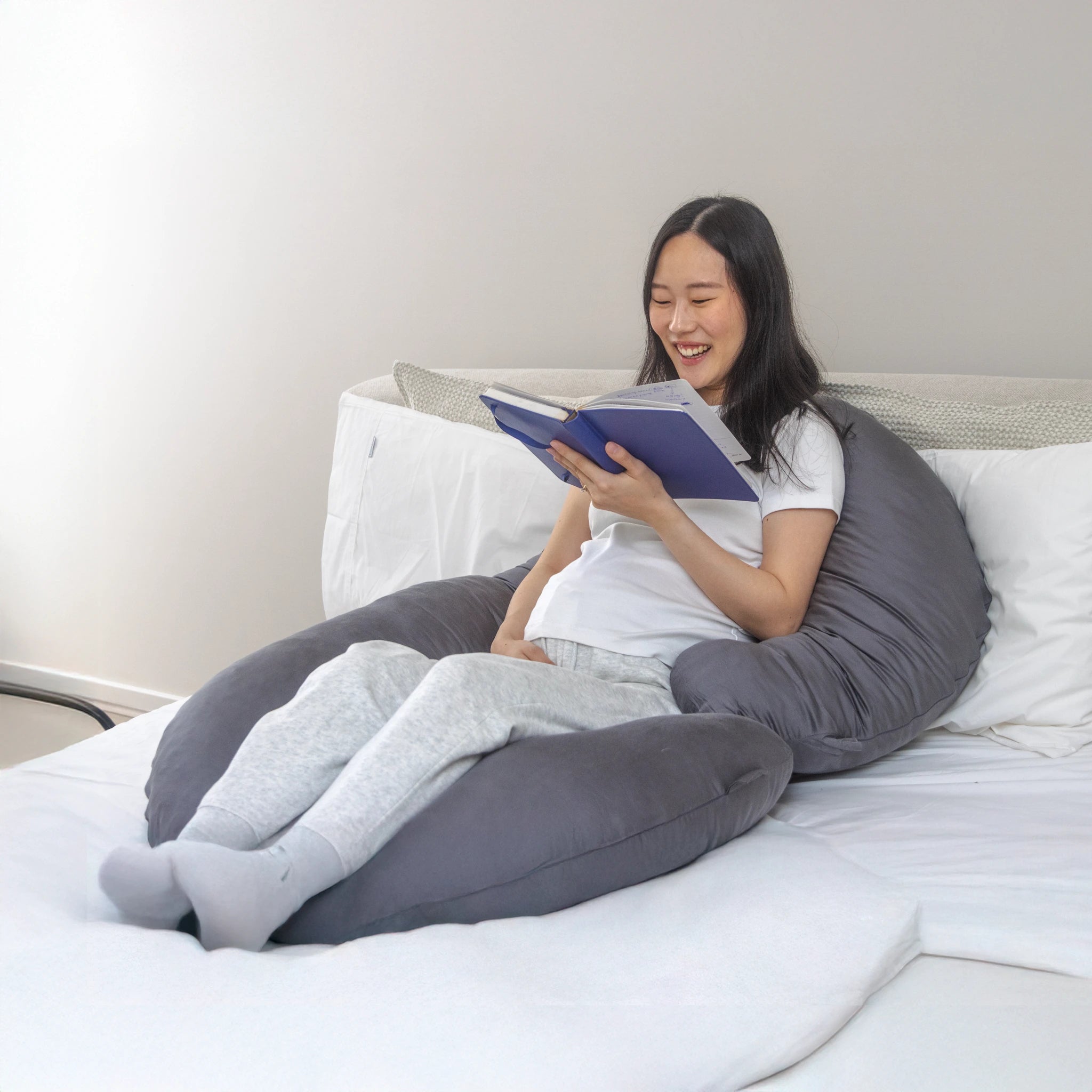 Pregnant woman smiling while reading with Comfybud C-shaped body and pregnancy pillow for back support, maternity comfort, and relaxation